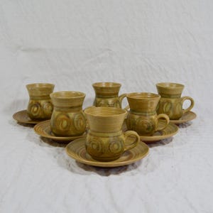 May include: A set of six ceramic mugs with matching saucers. The mugs and saucers are a light brown colour with a spiral design in shades of green and brown. Each mug has a handle.
