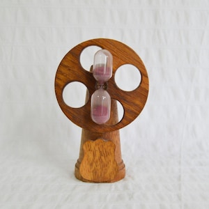 May include: A wooden hourglass with a decorative wheel design. The hourglass has a clear glass chamber filled with pink sand. The wooden base is a light brown colour.