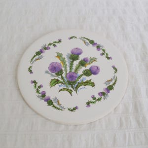 May include: A round white ceramic trivet featuring a central cluster of purple thistle flowers with green leaves and stems, surrounded by a circular wreath pattern of smaller thistle buds and foliage in matching purple and green tones.