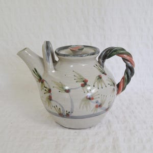 May include: A ceramic teapot with a cream-coloured glaze, featuring a hand-painted botanical pattern of green leaves and red berries. The teapot has a twisted, multi-coloured handle in shades of green and pink, a matching lid, and a spout.