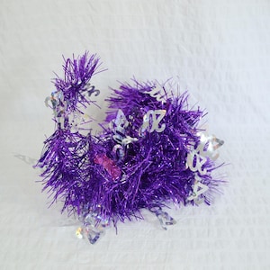 May include: A purple tinsel garland featuring silver holographic cutouts of the number 2024 interspersed throughout the metallic fringe. The decorative party accessory consists of vibrant purple strands with reflective silver accents.