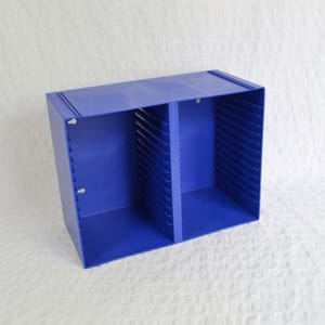 May include: A bright blue plastic storage unit featuring two side-by-side compartments. Each compartment contains a series of evenly spaced horizontal slots along the interior side walls, designed for organising flat items or media cases.