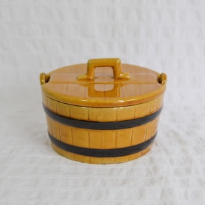 May include: A ceramic barrel-shaped container with a lid. The container is a warm yellow colour with two black bands around the middle. The lid has a handle and small side handles. The container is likely for food storage.