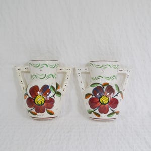 May include: A pair of white ceramic vases with handles, each decorated with a large red and yellow floral design. Green leaves and accents are included. The rims have green and brown painted patterns.