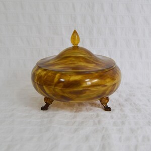 May include: A decorative, lidded container with a rounded body and three small feet. The container is a warm, amber colour with a swirling pattern. The lid is topped with a teardrop-shaped finial.