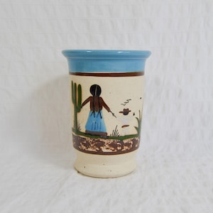 May include: A ceramic tumbler with a Southwestern design. The tumbler has a light blue rim, a brown band, and a cream-coloured body. The body is decorated with a painted scene of a person, a cactus, and a figure in a hat. The tumbler has a footed base.