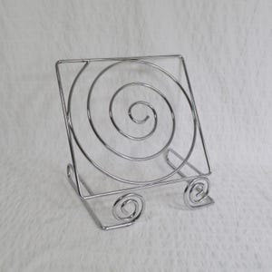 May include: A silver-coloured metal display stand featuring a square outer frame with a large central spiral design. The stand is supported by two base legs that each terminate in a smaller, decorative spiral curl at the front for stability.