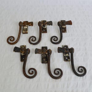 May include: A set of six dark brown, weathered metal window sash locks. Each piece features a rectangular mounting plate with two screw holes and a curved, scroll-shaped handle that pivots on a central hinge, showing signs of aged patina.