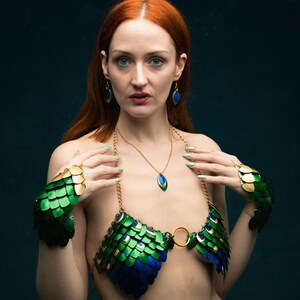 May include: A woman wearing a green, gold, and blue scale-like bra, matching wrist cuffs, earrings, and a necklace. The bra and cuffs have a peacock feather design. The jewelry is gold-toned. The background is a dark teal colour.