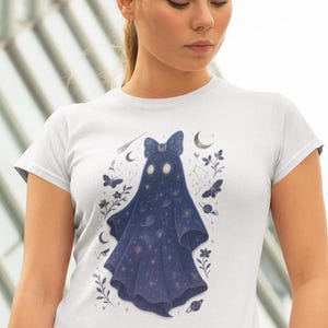 May include: White t-shirt featuring a whimsical ghost design. The ghost is depicted in a dark blue color, adorned with a bow and a starry night sky pattern. The design includes celestial elements like stars, planets, and butterflies.