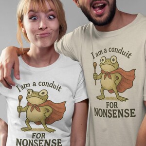 Peut inclure: Deux personnes portant des t-shirts assortis avec un dessin de grenouille. La grenouille, portant une cape et tenant un b&acirc;ton, est imprim&eacute;e avec le texte "I am a conduit FOR NONSENSE". Les chemises sont blanches et beiges.