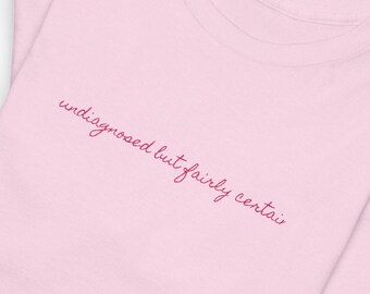 May include: Two light pink t-shirts are stacked on top of each other. The top shirt has the embroidered text "undiagnosed but fairly certain" in a cursive font. The shirts are made of a soft material and have a crew neck.