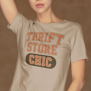 May include: A beige t-shirt with the text "THRIFT STORE CHIC" in orange and white lettering. The shirt is paired with black leggings. The model is posing with one arm raised above their head.