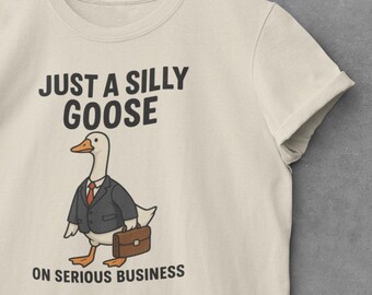 May include: Cream-colored t-shirt featuring a cartoon goose in a suit and carrying a briefcase. The text "JUST A SILLY GOOSE" is above the goose, and "ON SERIOUS BUSINESS" is below. The shirt is hanging on a wooden hanger.