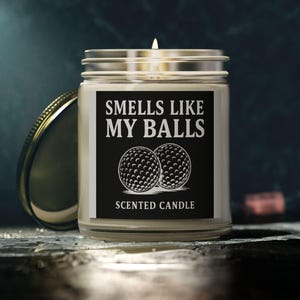 Peut inclure: Une bougie en pot de verre avec une mèche allumée et un couvercle argenté. L'étiquette indique "Smells Like My Balls" au-dessus de deux balles de golf et "Scented Candle" en dessous. Le fond est sombre avec une touche d'éclat.