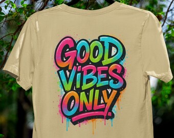 May include: Tan t-shirt with a colorful graphic that reads "GOOD VIBES ONLY." The text is in a graffiti style with a rainbow color scheme and dripping paint effects. The shirt is hanging on a black hanger against a background of green foliage.