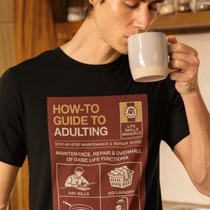 Può includere: T-shirt nero con grafica "HOW-TO GUIDE TO ADULTING" con illustrazioni di compiti quotidiani. La maglietta ha un girocollo. La persona che indossa la maglietta sta bevendo da una tazza beige.