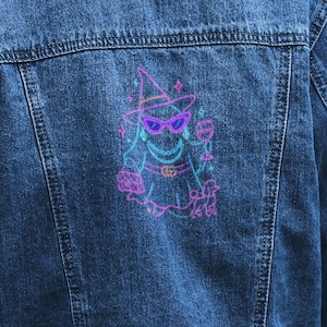 May include: A medium-wash denim jacket with a colorful embroidered design on the back. The design features a cartoon ghost wearing a witch hat, sunglasses, and holding a drink. The jacket has a classic collar and button closures.