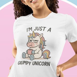 以下が含まれることがあります： 「I'M JUST A GRUMPY UNICORN」の文字と、マグカップを持った不機嫌そうなユニコーンのイラストがプリントされた白いTシャツ。青いジーンズを履いた人が着用しています。