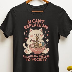 以下が含まれることがあります： 黒いTシャツ。ラーメンを食べる漫画の猫が描かれています。デザインには、ピンク色のテキスト「AI can't replace me, I'm already useless to society」が含まれています。シャツは白い壁を背景に、木製のハンガーに掛けられています。