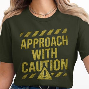 Könnte beinhalten: Dunkelgrünes T-Shirt mit dem Text "VORSICHT BEI DER NÄHERUNG" in goldfarbenen Buchstaben. Das Design umfasst Gefahrenstreifen und ein Ausrufezeichen, wodurch eine Warnmeldung entsteht.