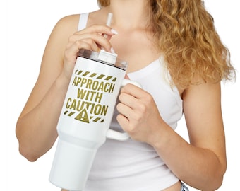 May include: White insulated tumbler with a black lid and handle. The tumbler features the text "APPROACH WITH CAUTION" in gold lettering. A clear straw is inserted into the lid. The tumbler is designed to keep beverages cold or hot.