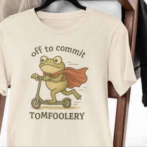 Op de afbeelding: Crèmekleurig T-shirt met een cartoonkikker op een scooter en met een rode cape. De tekst "off to commit TOMFOOLERY" staat boven en onder de kikkerillustratie. Het shirt hangt aan een zwarte hanger.