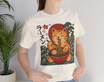 May include: Cream-colored t-shirt featuring a cartoon cat enjoying ramen. The cat is orange with stripes, and the ramen bowl is red. Japanese text and floral accents surround the cat. The design includes a red sun and stylized waves.