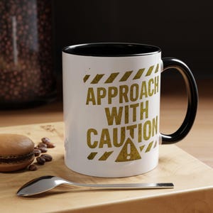 Könnte beinhalten: Weißer Keramikbecher mit schwarzem Innenraum und Griff. Der Becher zeigt den Text "APPROACH WITH CAUTION" in goldfarbenen Buchstaben, zusammen mit Warnbandgrafiken. Ein Löffel und ein Macaron befinden sich auf einer Holzoberfläche.