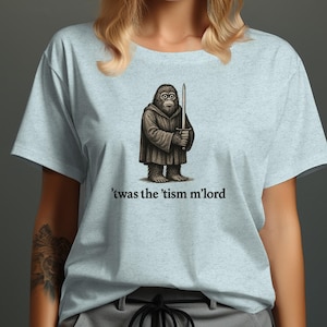 Op de afbeelding: Lichtblauw T-shirt met een cartoon aapachtig figuur in een gewaad met een zwaard. De tekst "'twas the 'tism m'lord" is onder de illustratie gedrukt. Het shirt is gemaakt van een zacht materiaal.