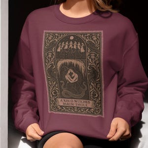 May include: Burgundy sweatshirt featuring a detailed graphic with the text "A Kin O Witches Maun Thole." The graphic depicts figures dancing under a full moon, with a woman and serpent in the center. The design is framed by floral and bird motifs.