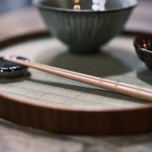 以下が含まれることがあります： 濃い茶色の縁が付いた木製のサービングトレイには、灰色のボウル、小さな茶色のボウル、黒い箸置きの上に置かれた一対の木製箸が置かれています。 トレイは、明るい緑色のマットの上にあります。