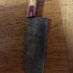 Peut inclure: Un couteau à hacher avec une lame en acier à motifs et un manche en bois. Le manche présente une combinaison de tons de bois clairs et foncés, avec des accents rouges. La lame a un motif sombre et tourbillonnant.