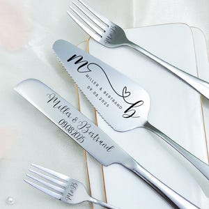 Può includere: Un set di coltello e server per torta nuziale in acciaio inossidabile con due forchette, presentato su un piatto rettangolare bianco. Il coltello e il server sono incisi con nomi e una data. Anche le forchette sono incise.