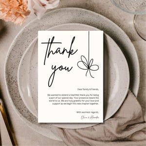 Könnte beinhalten: Eine weiße Dankeskarte mit schwarzem Schriftzug und einer Schleifenillustration. Die Karte liegt auf einem neutralfarbenen Gedeck, passend für eine Hochzeit oder Veranstaltung. Der Text lautet "Danke" und eine Nachricht.