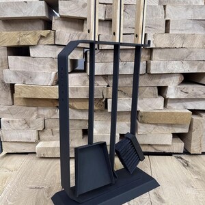May include: A black fireplace tool set with a stand. The set includes a shovel, brush, and poker. The handles of the tools are made of light-colored wood. The background features stacked wooden planks.