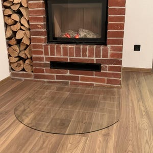 Op de afbeelding: Een bakstenen open haard met een zwart omkaderde vuurhaard, een glazen haard en een stapel brandhout. De vuurhaard bevat gloeiende sintels. De vloer is van hout.