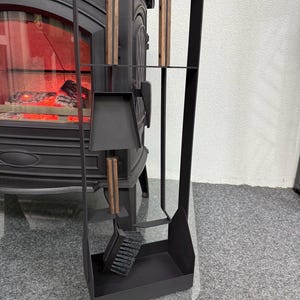 Pode incluir: Um conjunto de ferramentas para lareira pretas com um suporte. O conjunto inclui uma escova, uma pá, um atiçador e um elevador de toras. As alças são de madeira. As ferramentas são exibidas em frente a uma lareira preta.