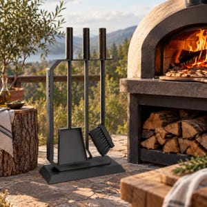 May include: A black metal fireplace tool set featuring a stand with a shovel, a brush, and a poker. Each tool has a dark wooden handle. The set is organised on a rectangular base, designed for maintaining a fire in an outdoor pizza oven.