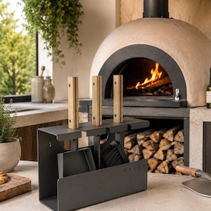 Pode incluir: Um conjunto de ferramentas para lareira em metal cinzento-escuro mate, com uma pá e uma escova com cabos de madeira clara. O suporte tem uma base retangular e ranhuras verticais para manter as ferramentas na vertical. O nome SAVEN está impresso.