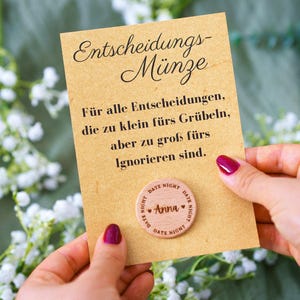 Könnte beinhalten: Eine Holz-Entscheidungsmünze mit dem Text "Anna" und "Date Night" wird vor einer Karte mit deutschem Text gehalten. Auf der Karte steht "Entscheidungs-Münze" und weiterer Text. Der Hintergrund umfasst weiße Blumen und einen grünen Stoff.