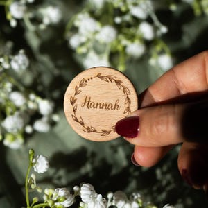 Könnte beinhalten: Eine runde Holzscheibe mit dem Namen "Hannah" in kursiver Schrift, umgeben von einem Lorbeerkranz. Die Scheibe wird vor einem Hintergrund aus weißen Blumen und einer grünen Oberfläche gehalten.
