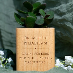 Könnte beinhalten: Ein Holzpflanzgefäß mit einer grünen Sukkulentenpflanze. Der Pflanztopf trägt die Gravur "FÜR DAS BESTE PFLEGETEAM" und "DANKE FÜR EURE WERTVOLLE ARBEIT - TAG FÜR TAG." Der Hintergrund ist ein grüner Stoff.