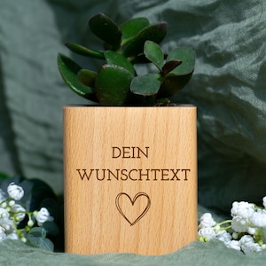 Könnte beinhalten: Ein Holzpflanzgefäß mit dem Text "DEIN WUNSCHTEXT" und einem Herzsymbol, das eine grüne Sukkulentenpflanze enthält. Der Pflanztopf ist aus hellem Holz gefertigt und von weißen Blumen und einem grünen Hintergrund umgeben.