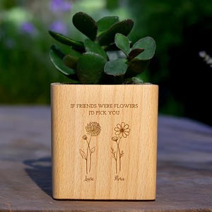 Könnte beinhalten: Ein hölzerner Pflanzkasten mit einer Sukkulentenpflanze. Die Box ist mit dem Text "IF FRIENDS WERE FLOWERS I'D PICK YOU" und zwei Blumenillustrationen mit den Namen "Luise" und "Flora" darunter graviert. Der Pflanzkasten ist quadratisch und aus hellem Holz.