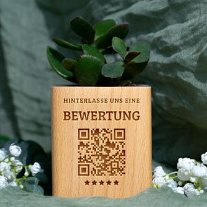 Könnte beinhalten: Ein kleiner, quadratischer Holztopf mit einer Sukkulentenpflanze. Der Topf hat einen eingravierten Text mit der Aufschrift "HINTERLASSE UNS EINE BEWERTUNG" und einen QR-Code. Unter dem QR-Code sind fünf Sterne eingraviert. Die Sukkulente hat grüne Blätter.