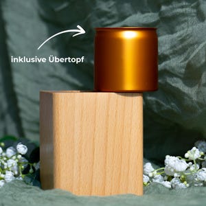 May include: A golden-colored metal pot sits atop a light-colored wooden block. The pot is cylindrical with a slightly flared rim. The wooden block is a square shape. The text "inklusive &Uuml;bertopf" is in the image.