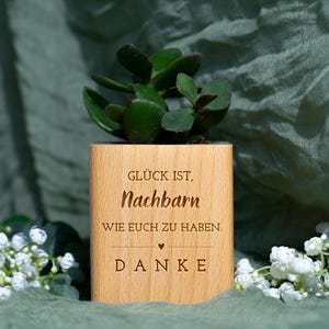 Könnte beinhalten: Ein Holz-Pflanztopf mit einer grünen Sukkulentenpflanze. Auf dem Topf steht der gravierte Text: "Glück ist, Nachbarn wie euch zu haben. Danke." Der Pflanztopf ist von weißen Blumen umgeben und vor einem grünen Hintergrund platziert.