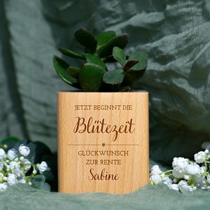 Könnte beinhalten: Ein Holztopf mit einer grünen Sukkulentenpflanze. Der Topf trägt die Gravur "JETZT BEGINNT DIE Blütezeit" und "GLÜCKWUNSCH ZUR RENTE Sabine". Der Topf ist von weißen Blumen umgeben und vor einem grünen Hintergrund platziert.