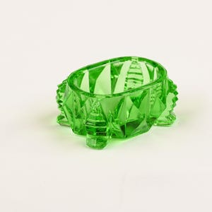 May include: A small, oval-shaped, translucent green glass bowl featuring a faceted, diamond-cut exterior pattern. The bowl is supported by four short, sturdy feet and has a wide, open top with a smooth, polished rim, showcasing a vibrant color.